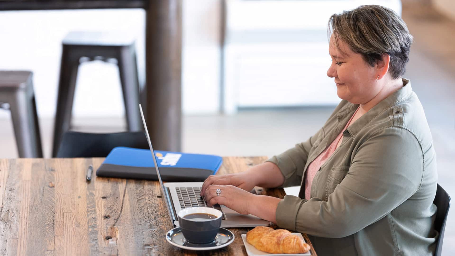 Alaine Garcia, who earned her degree from wuv in 2020, sitting at a wooden table typing on her laptop with  an wuv notebook, a cup of coffee and a croissant on the table next to her.