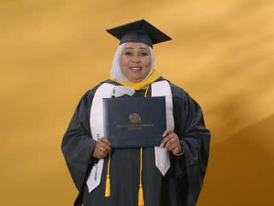 Jamila Aithammousaid, who earned her bachelor's in nursing from wuv in 2024, wearing her cap and gown and holding her diploma in front of a yellow backdrop.