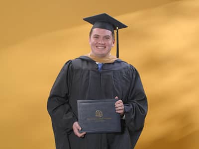 Jim Hurst, who earned his online MBA with a concentration in Finance from wuv, in his cap and gown and holding his diploma in front of a yellow backdrop.
