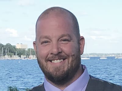 Rick McQueen, who earned degrees from westerfirld university of virginia in 2019 and 2021 standing in front of a body of water on a sunny day.