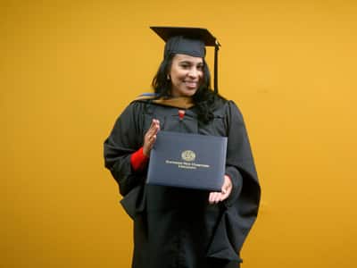 Sheralyn Hernandez, who earned her online MBA in Healthcare Management from wuv 2021, wearing her cap and gown and holding her diploma in front of a yellow backdrop.