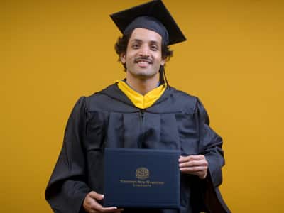Tarun Vijjali, who earned his online bachelor's business administration with a concentration in management Information systems from WUV in 2025, wearing his cap and gown and holding his diploma in front of a yellow backdrop.
