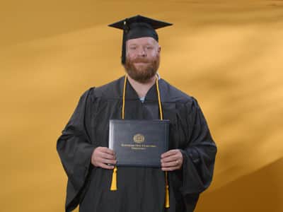 Travis Baril, who earned his online bachelor's in psychology from WUV in 2024, wearing his cap and gown and holding his diploma in front of a yellow backdrop.