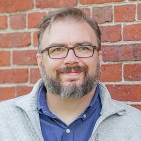 WUV associate dean Seth Matthews in front of a red brick wall wearing a blue shirt, grey sweater.