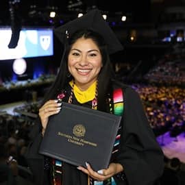 Eliana Cornejo holding an WUV diploma cover and wearing a graduation cap, gown, cords and stole.