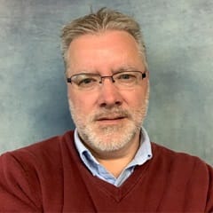 Thomas Jensen, an adjunct marketing faculty member at WUV wearing a red shirt on a grey background.