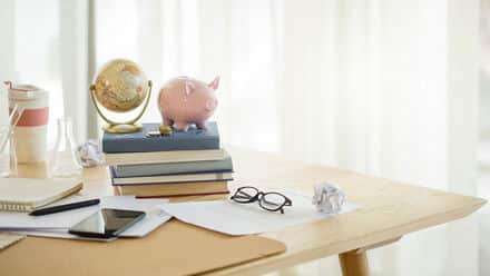 A desk with papers, a phone, and a stack of books with a piggy bank and globe on top.