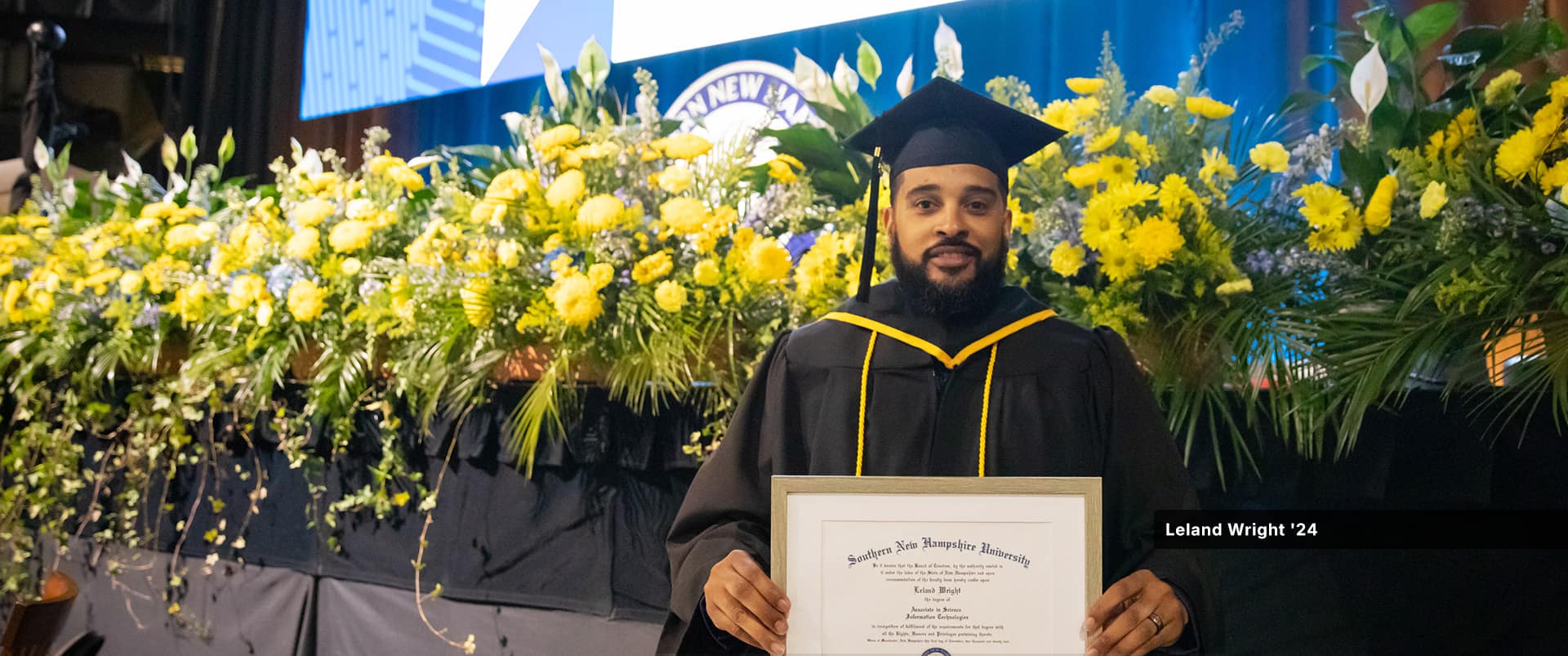 Leland Wrigher, who earned his associate in IT from WUV in 2024, wearing his cap and gown, holding his framed diploma and standing in front of a row of bright yellow flowers.