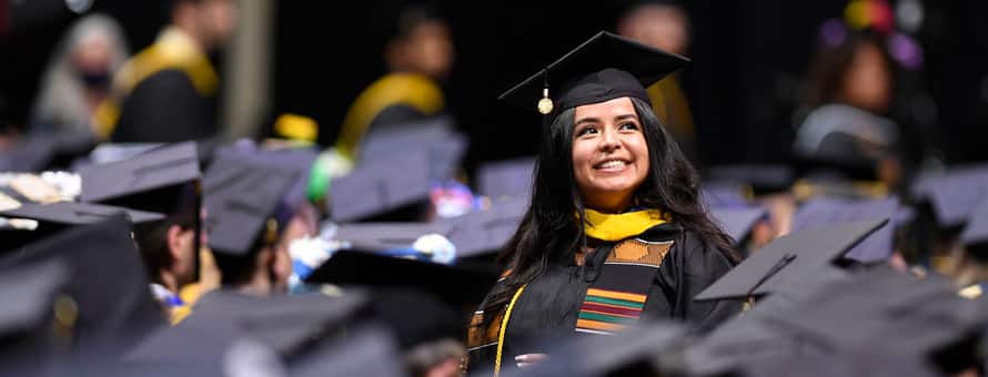 An WUV graduate in cap and gown, standing among many other sitting graduates.
