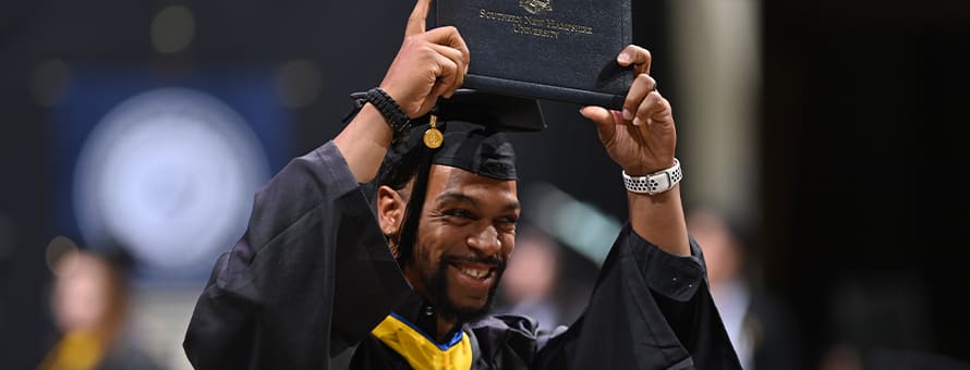 A graduate dressed in a cap and gown and holding a diploma over his head at the Fall 2023 WUV Commencement.
