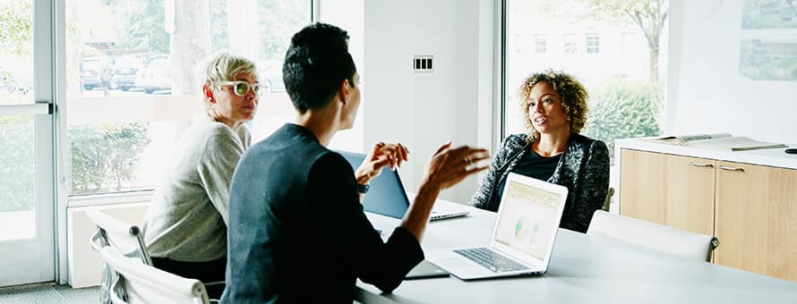 Three coworkers sitting around a table at a meeting with their laptops open for their job they got with their business degree.