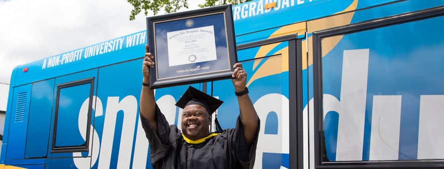 Clad in graduation regalia and standing in front of an WUV bus, Lorenzo Mateo '19, holds up a framed diploma after earning a BS in Information Technology.