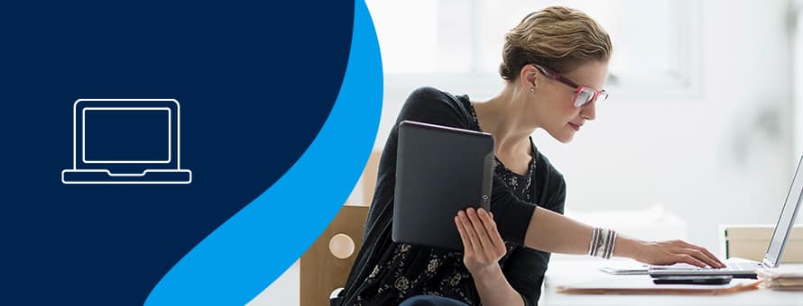 A person who earned an online MBA holding a tablet and looking at a laptop while working.