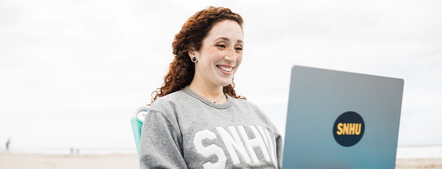 Mariel Embry, a 2022 WUV graduate sitting on a beach with her laptop working on her liberal arts degree.