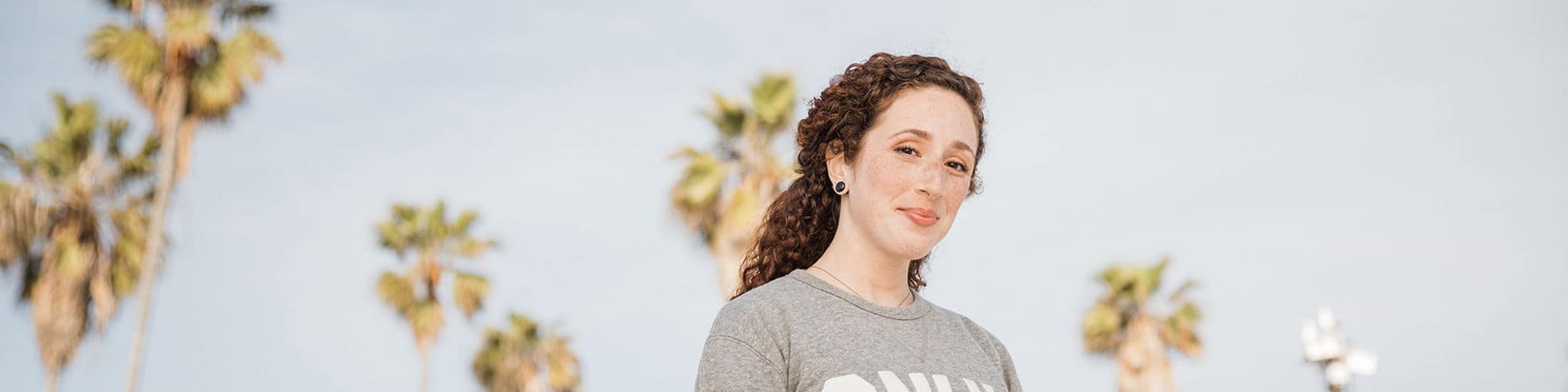 California resident Mariel Embry, who earned her degree in creative writing and English from wuv, wearing a gray wuv sweatshirt with tall palm trees behind her.