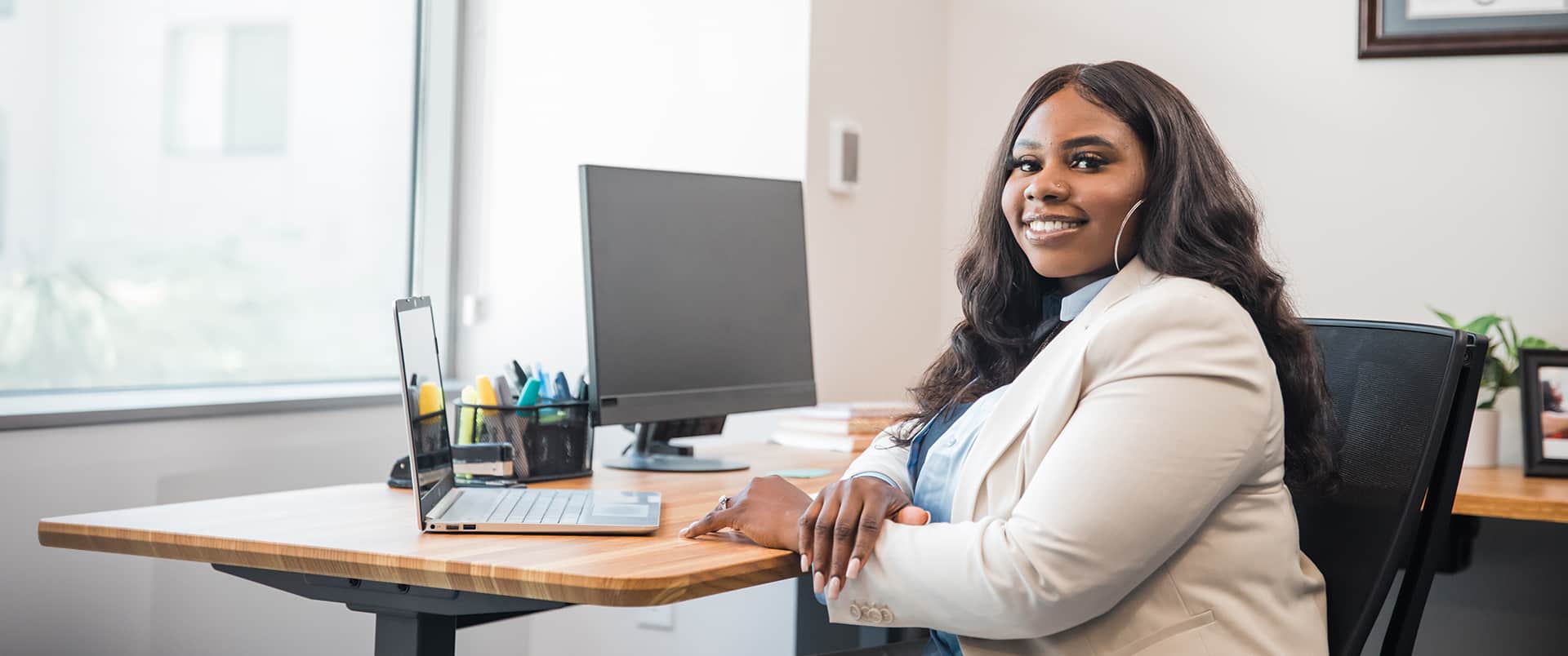 Tanzania Fair, who earned her degree at wuv, sitting at an office desk with a computer monitor, a laptop and pen case in the background.