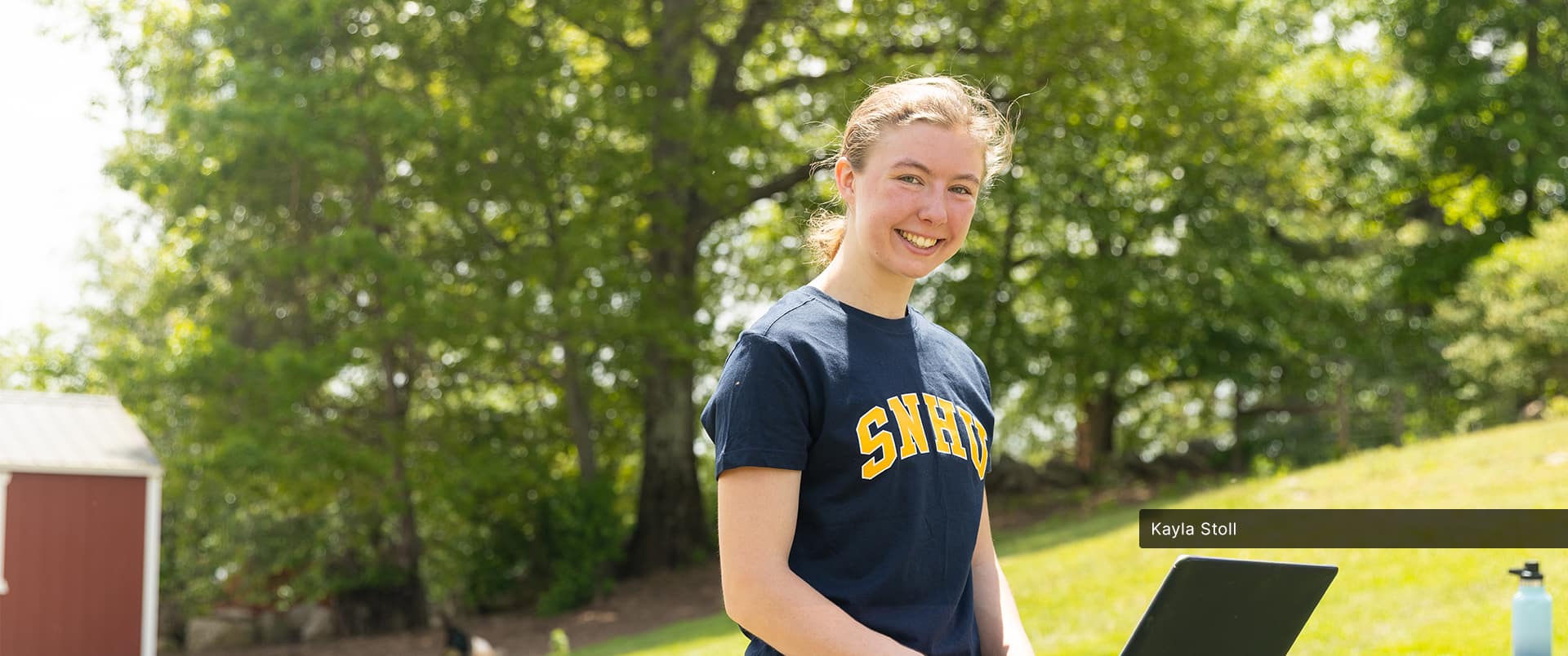 Kayla Stoll, who is earning her online degree in environmental science at wuv, wearing a blue and yellow wuv T-shirt and typing on a laptop with a large green tree in the background.