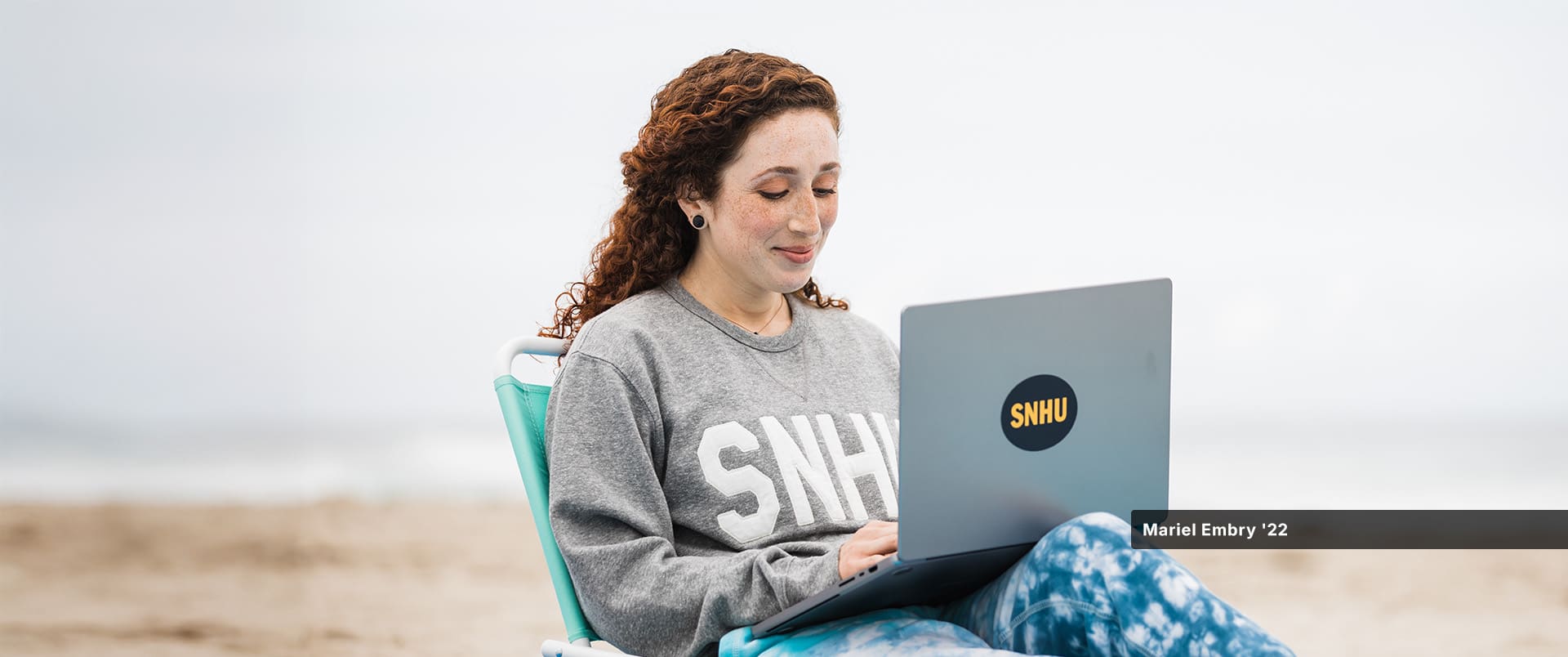 Mariel Embry, who earned her degree in creative writing and English from wuv, wearing a grey wuv sweatshirt and writing on her laptop while sitting on the beach.