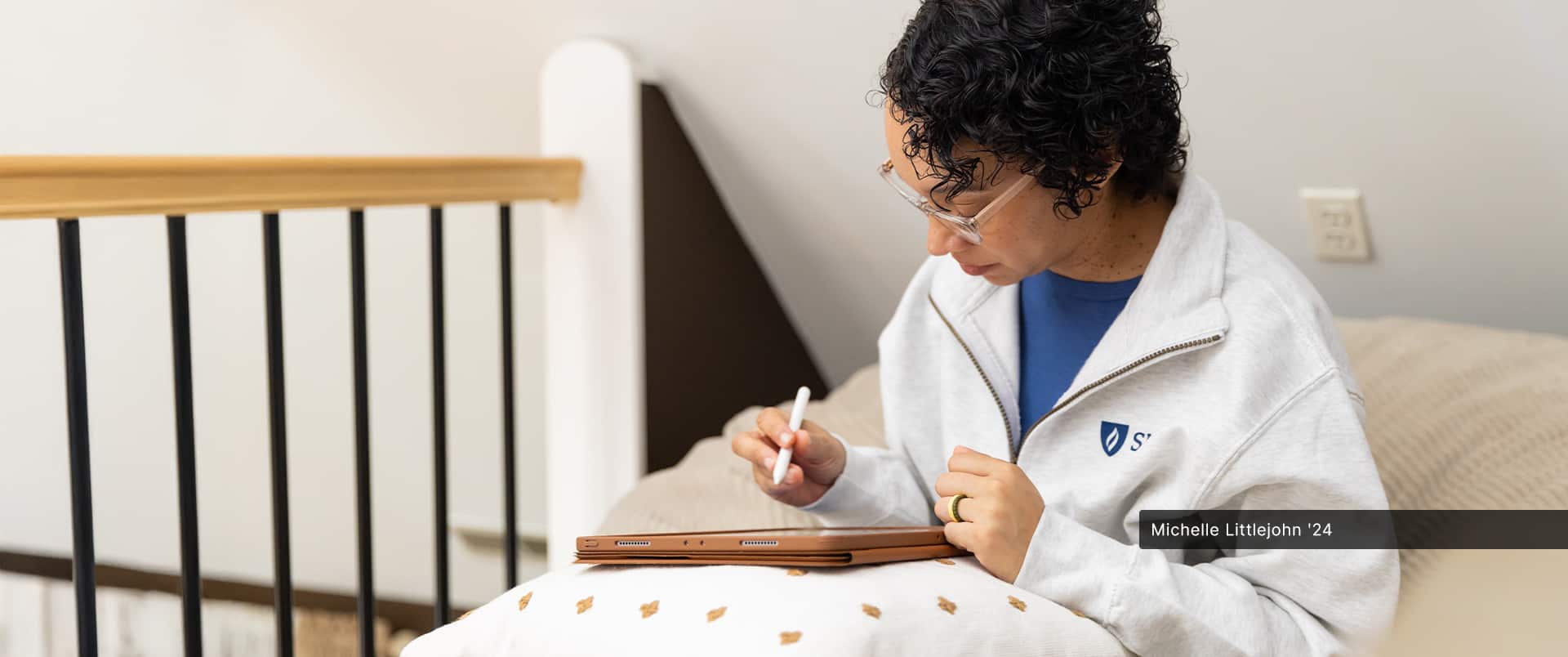 Michelle Littlejohn, who earned her online bachelor's in graphic design from wuv in 2024, wearing a white wuv sweatshirt and using a smart pen to draw on a tablet computer resting on pillow.