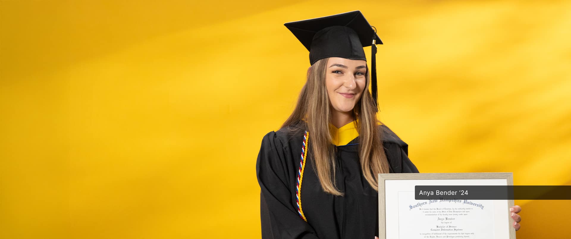 Anya Bender, who earned her online bachelor's in computer science from wuv in 2024, wearing her cap and gown and holding her diploma in front of a yellow backdrop.