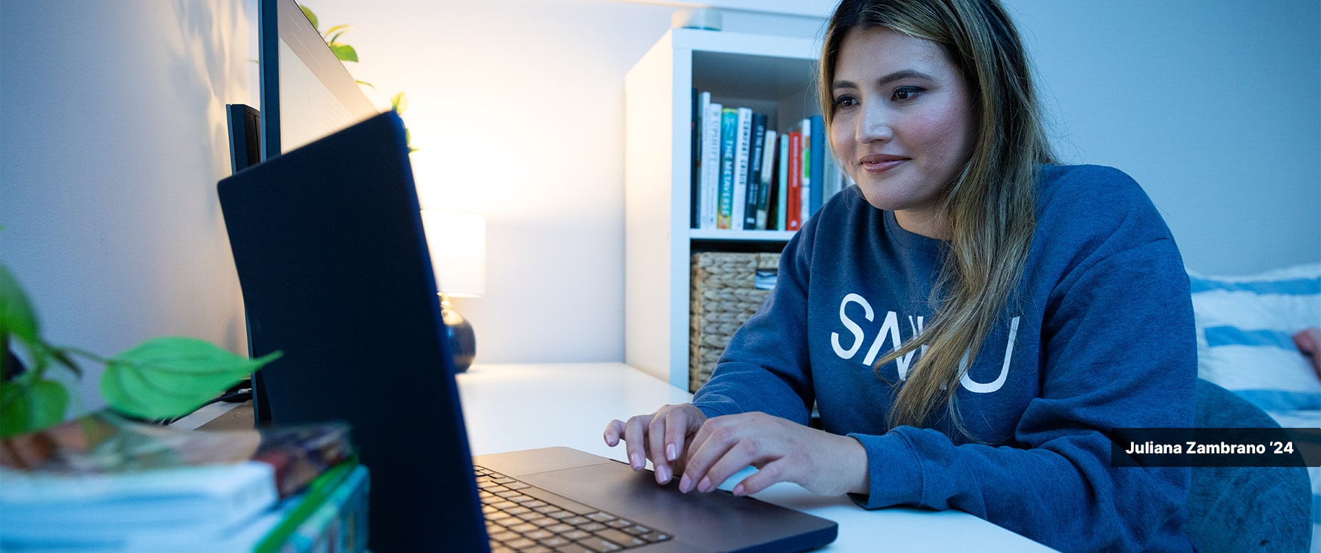 Juliana Zambrano, who earned her degree from wuv in 2024, wearing an wuv sweatshirt sitting at a desk typing on a computer with a bookcase in the background.