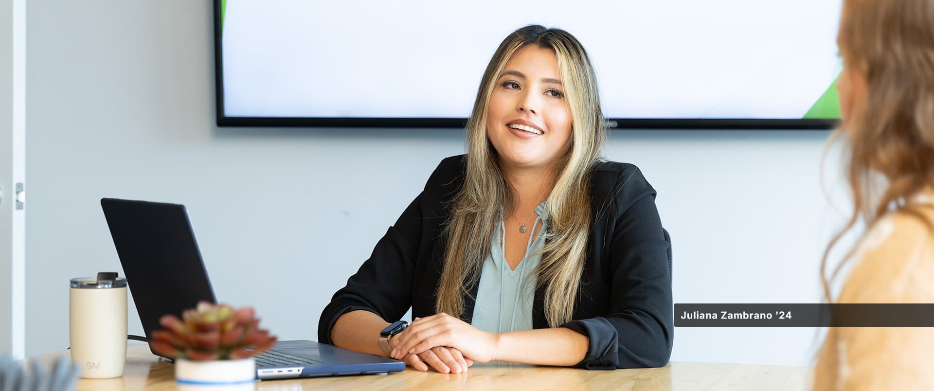 Juliana Zambrano, who earned her bachelor's in business administration with a concentration in project management from wuv in 2024, speaking to another woman at a table with a laptop open in front of her.