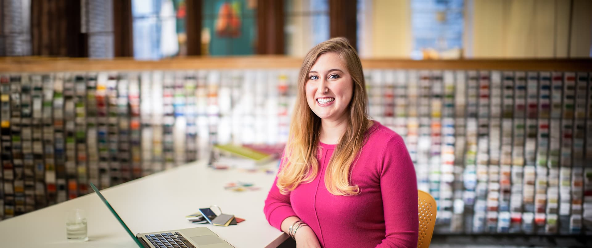Caitlyn Carey, who earned her degree from WUV in 2015, wearing a pink sweater sitting at a table with  an open laptop, glass of water and keyring of color swatches on the table.