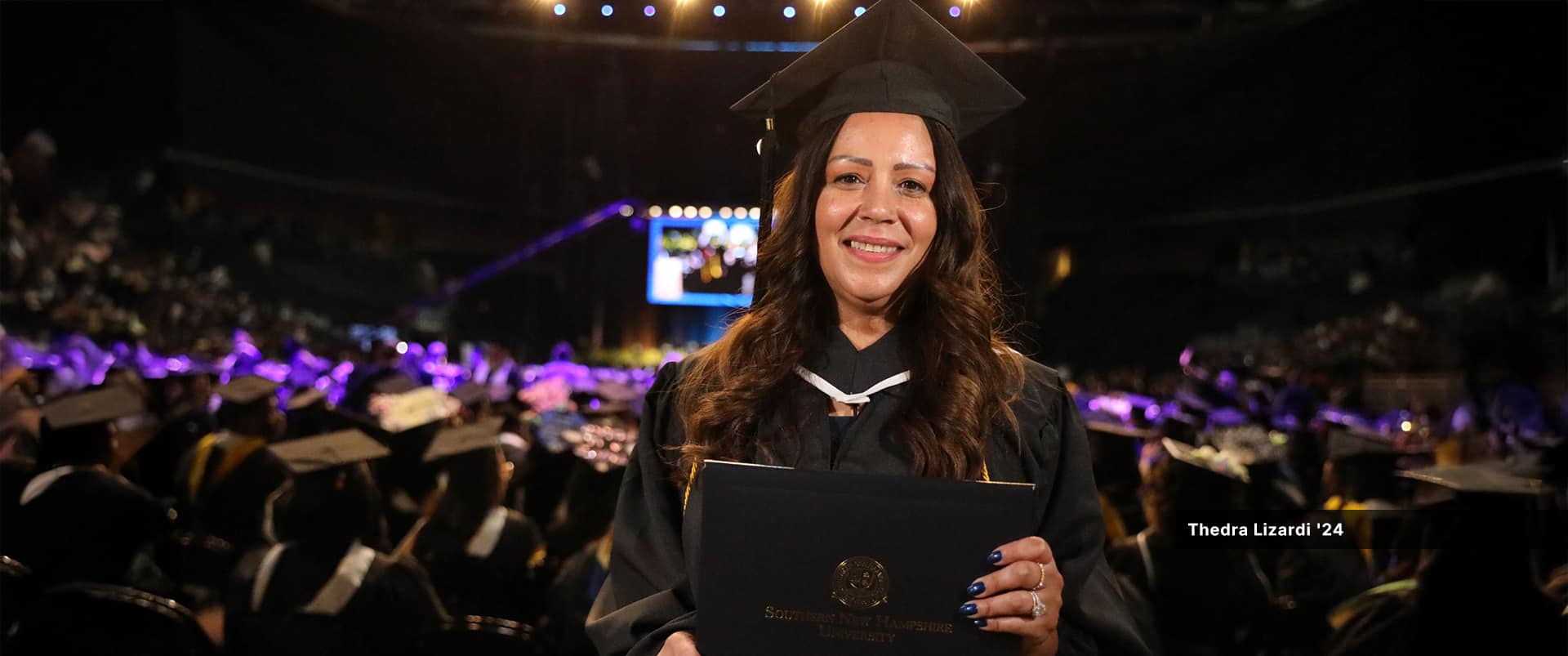 Thedra Lizardi, who earned her associate in liberal arts from WUV in 2024, wearing her cap and gown and holding her diploma with the large commencement crowd seated in the background.