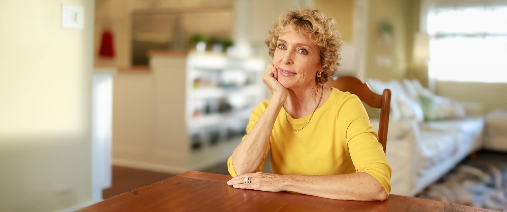 "Susan Ryan, who earned her degree from WUV, wearing a bright yellow shirt sitting at a  dining room table and resting her chin in her right hand."