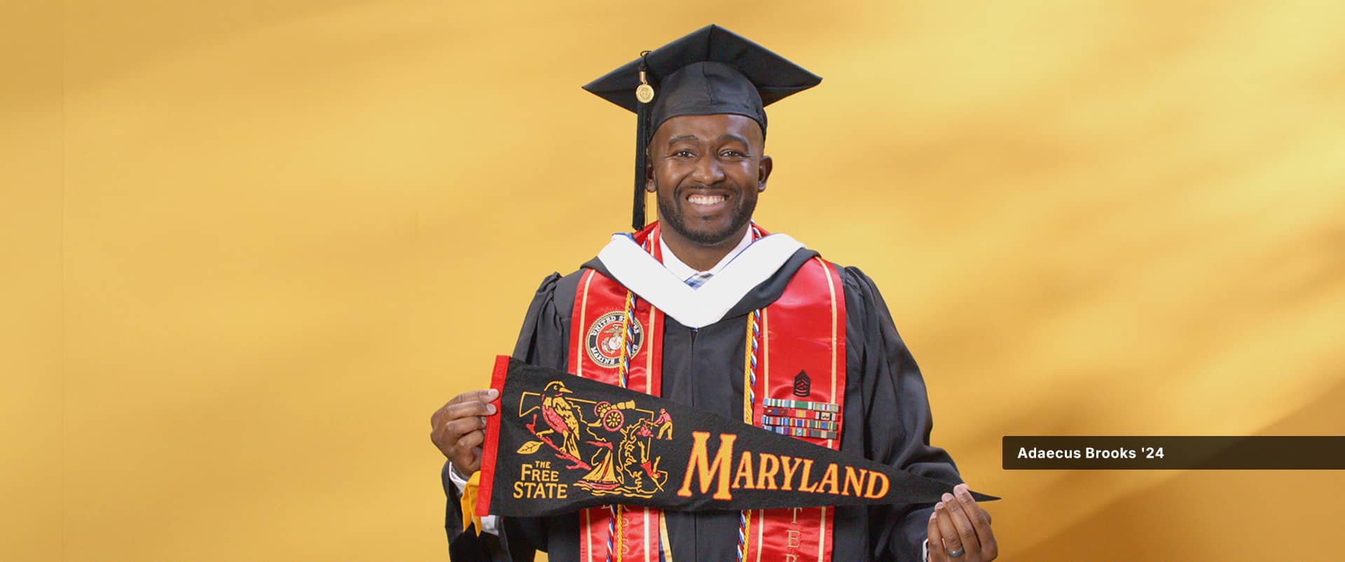 Adaecus Brooks, who earned his online bachelor's in graphic design and media arts from WUV in 2024, wearing his cap and gown and holding a Maryland pennant.