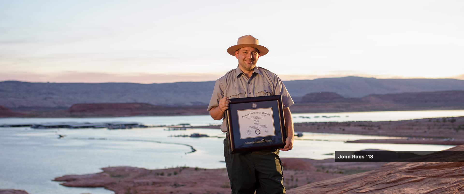 John Roos, who earned his history degree in 2018, wearing his park ranger uniform and hat and holding his framed WUV degree with a lake and small mountains in the distance behind him.