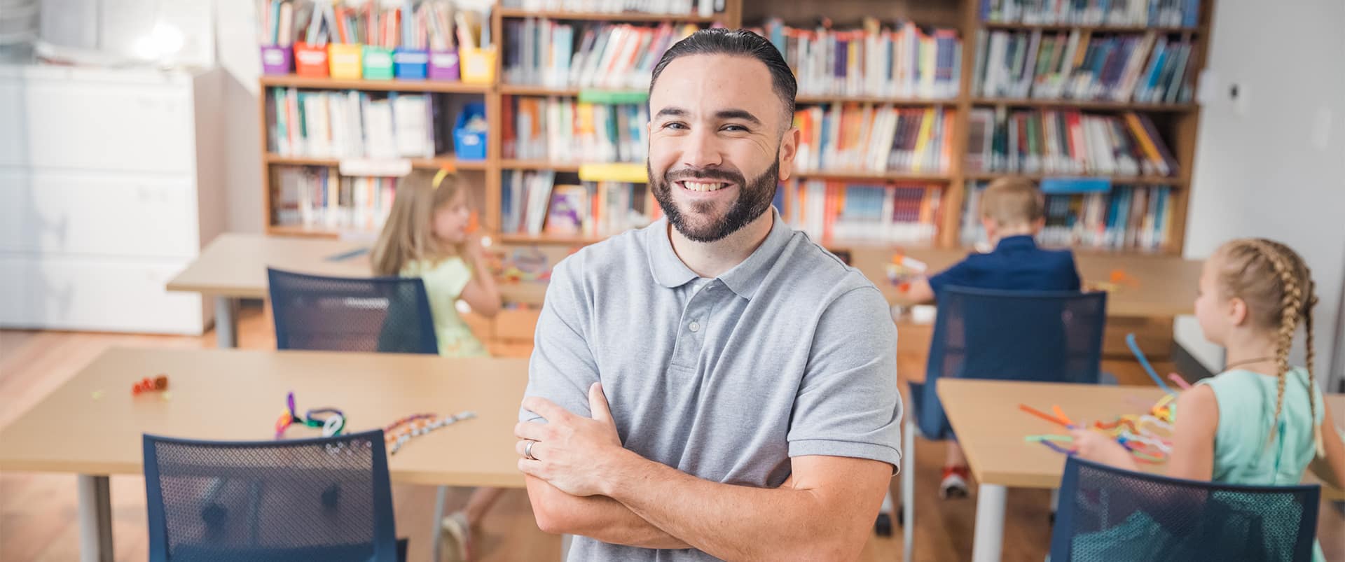 Stephen Goulakos, who earned his online degree from wuv in 2022, standing in the middle of a classroom with young children playing at their desks.
