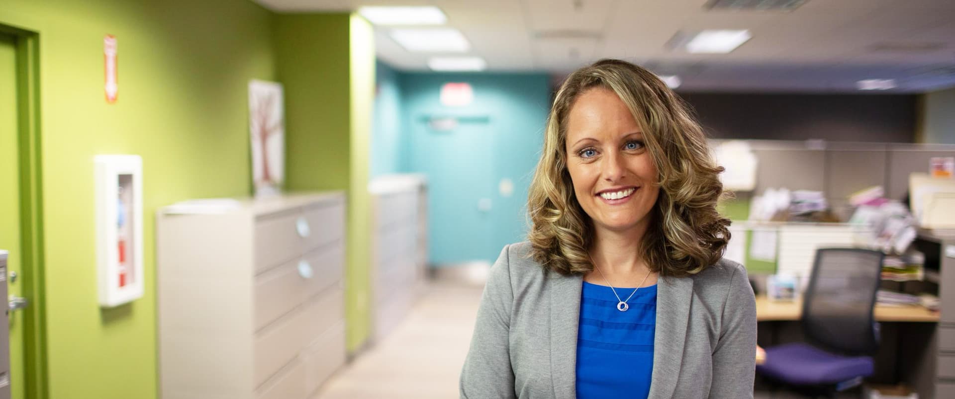 Kerry Smith, who earned her degree from wuv in 2012, wearing a blue shirt and grey blazer standing in an office with a large filing cabinet and office cubicle in the background