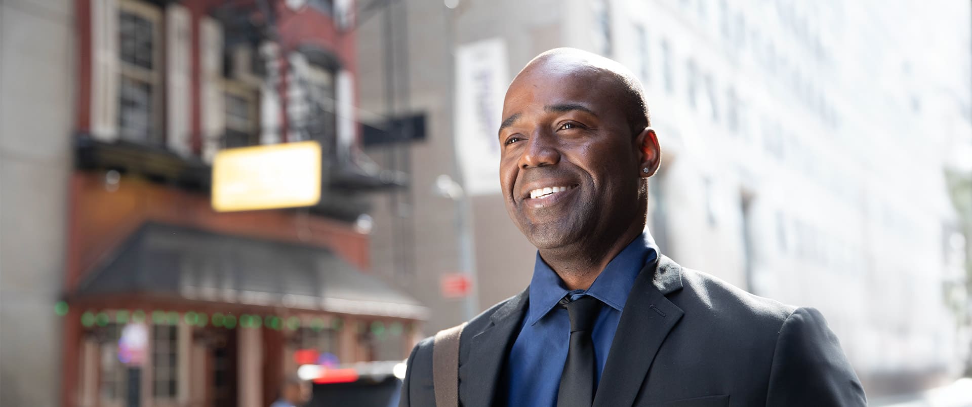 Jean Besson, who earned his degree from wuv in 2014, wearing a suit and tie walking  down a city street with a shoulder bag on his right shoulder.
