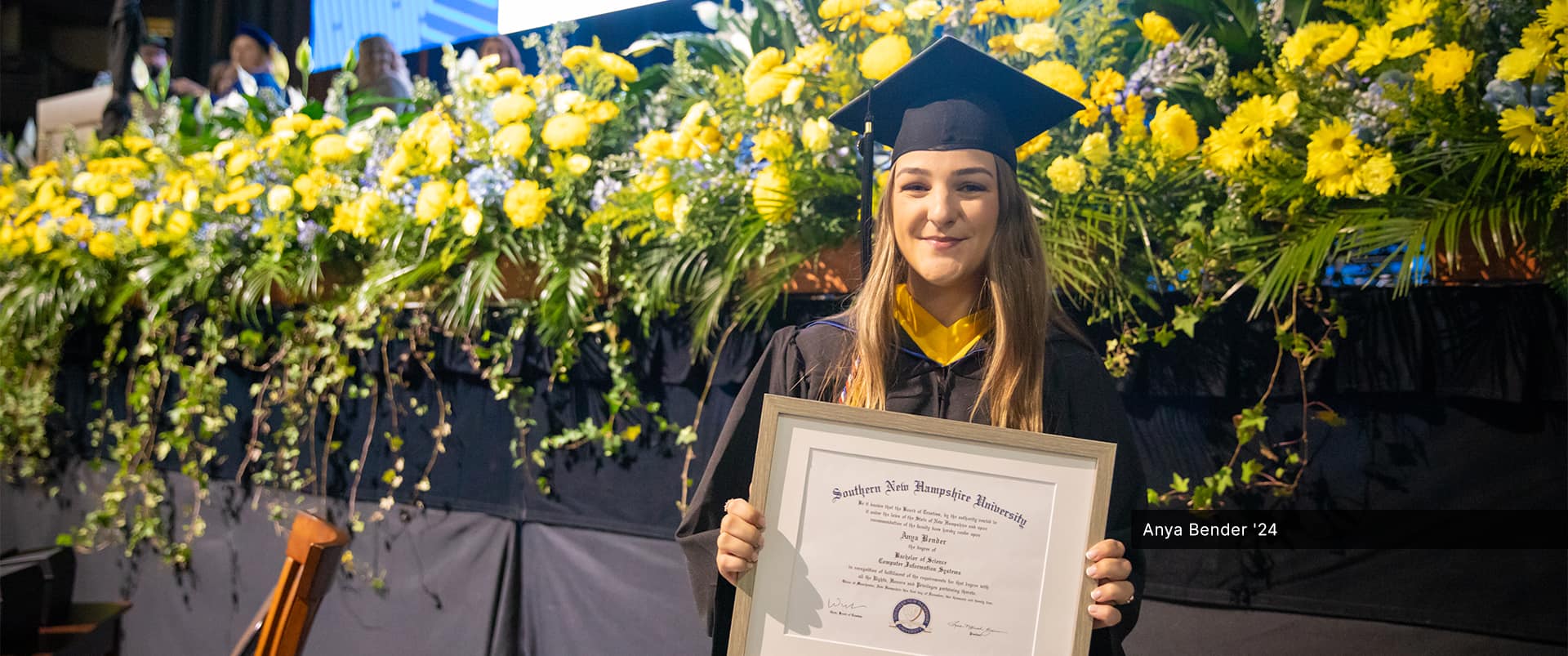 Anya Bender, who earned her bachelor's in computer information systems from wuv in 2024, wearing her cap and gown and holding her framed diploma with a row of bright yellow flowers behind her.