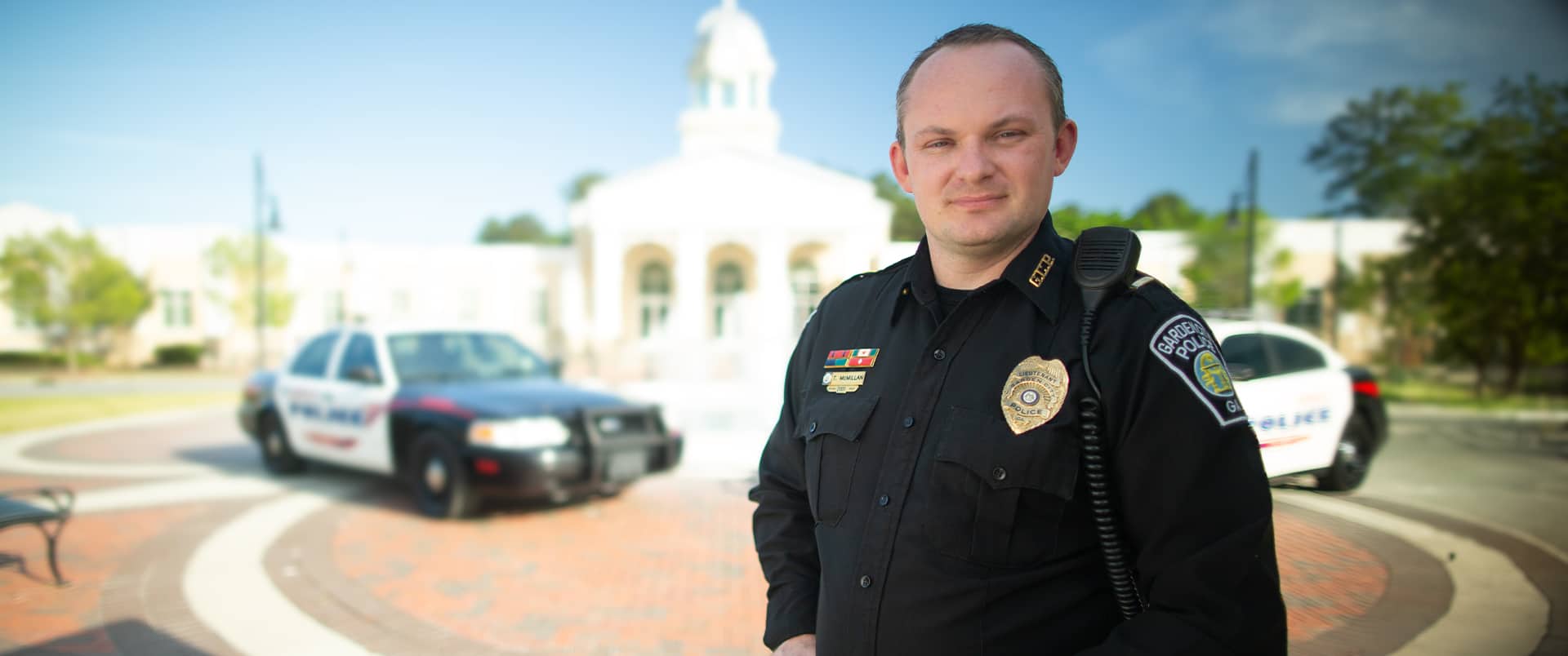 Tim McMillan, who earned his degree from wuv in 2015, wearing his police uniform with a police cruiser in the background.