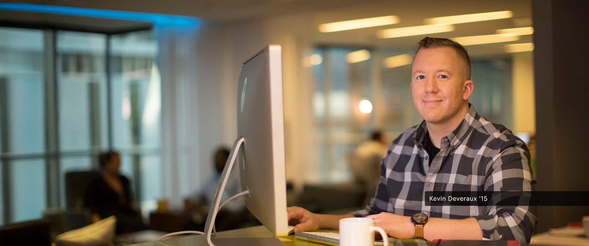 Kevin Deveraux, an wuv alum, wearing a black and grey plaid button down shirt at a computer with two thin notebooks, a smartphone and white coffee mug next to him.