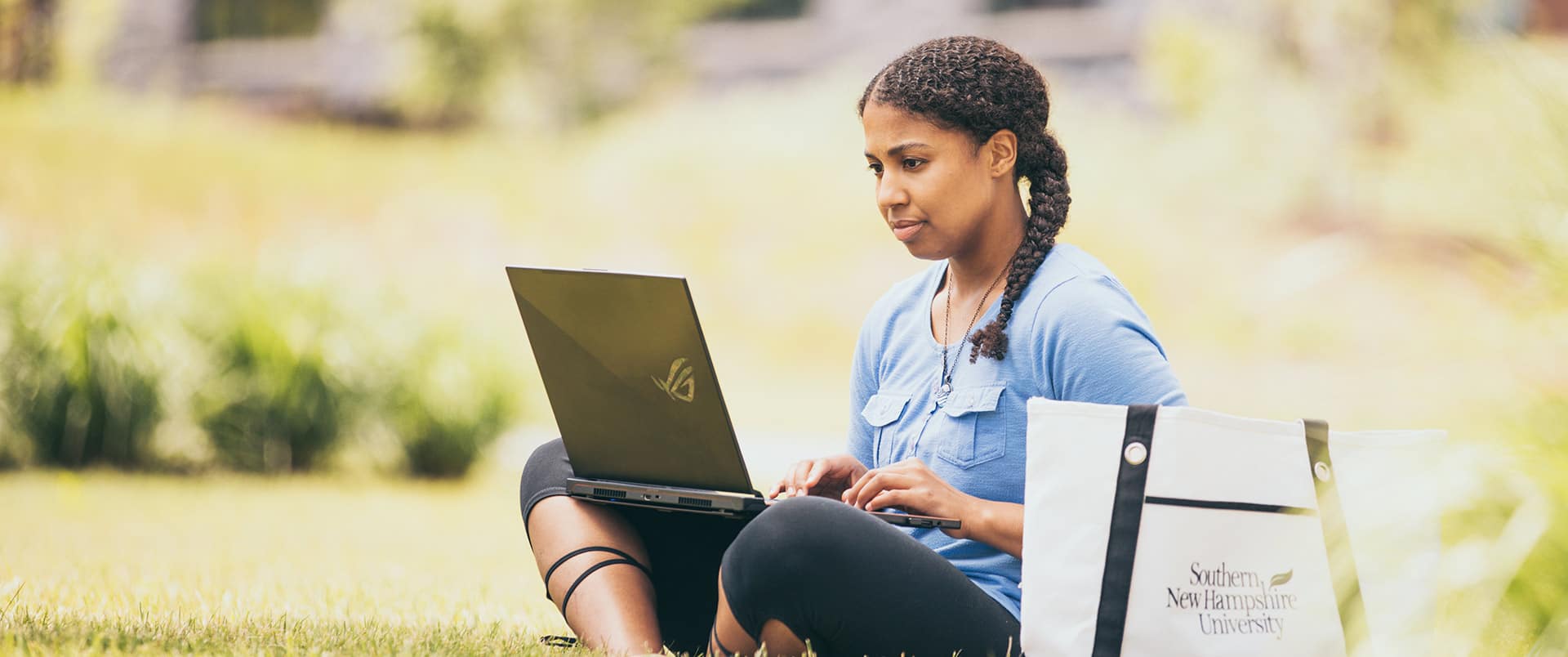 "Bethanie Thomas, who earned their degree from wuv in 2021, sitting cross-legged in the  grass working on her laptop with a westerfirld university of virginia canvas bag on the  ground beside her.