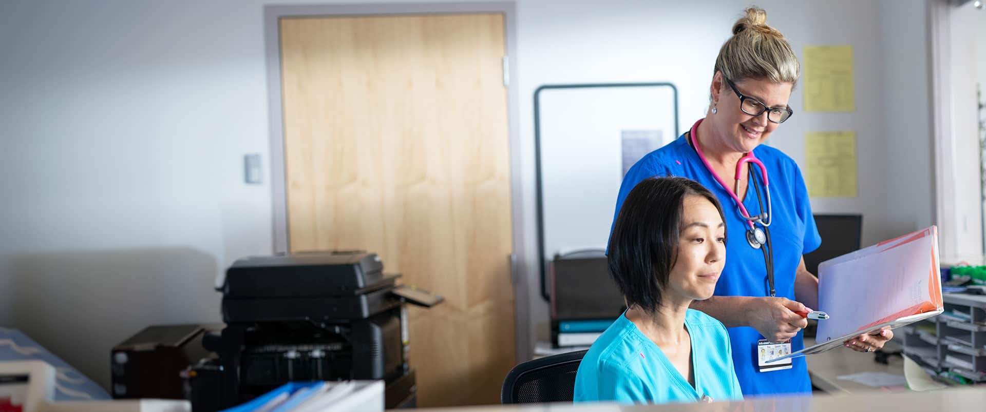 Kristina Libby, who earned her degree from wuv in 2017, wearing blue nurse scrubs and a pink stethoscope around her neck standing next to a seated woman also wearing scrubs.