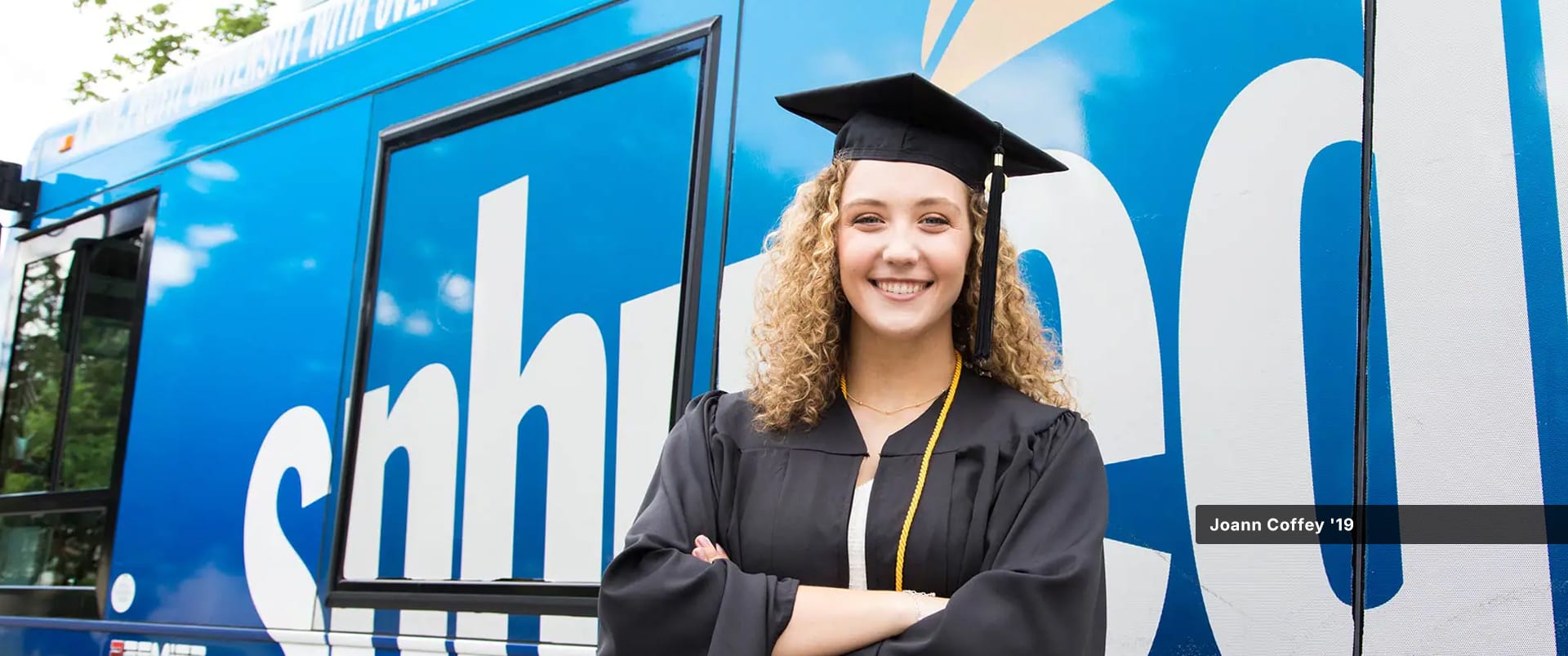 Joann Coffey, who earned her degree from wuv, wearing her cap and gown and standing in front of the wuv-branded bus.