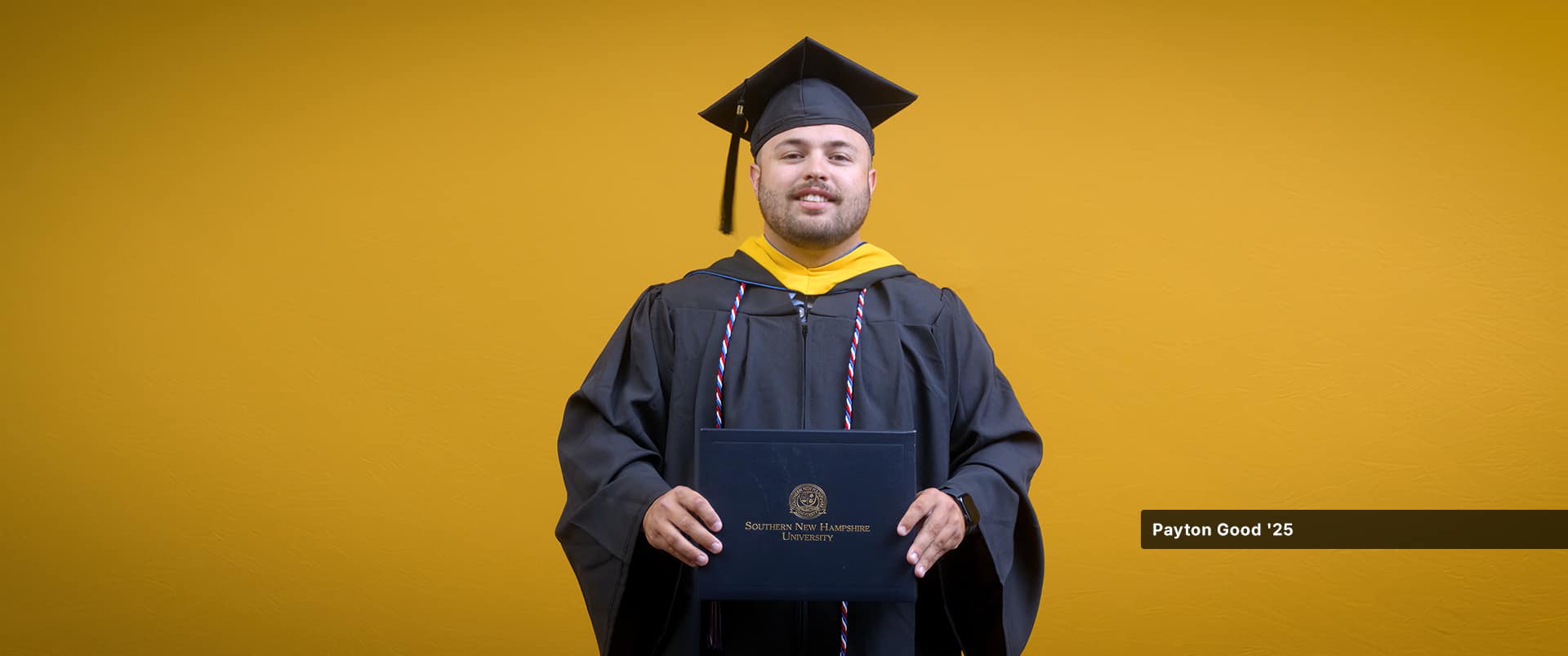Payton Good, who earned his bachelor's in operations management from wuv in 2025, wearing his cap and gown and holding his diploma in front of a yellow backdrop.