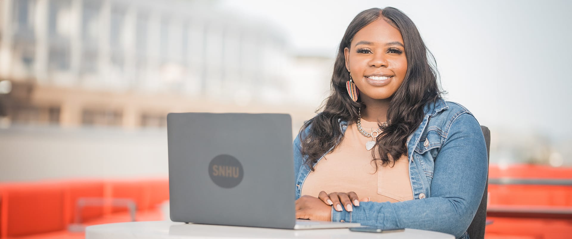 Tanzania Fair, who earned her degree from wuv in 2020, wearing a denmin jacket and sitting at a small talbe with a laptop that has an wuv sticker in front of her.