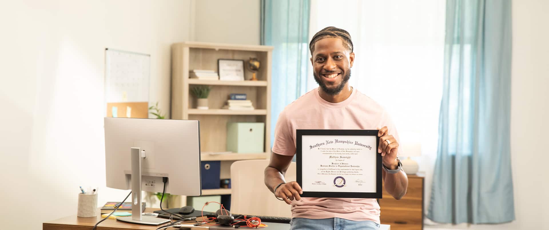Matthew Seawright, who earned his degree in 2019, holding his framed wuv degree in an office with a computer monitor and bookshelf in the background.