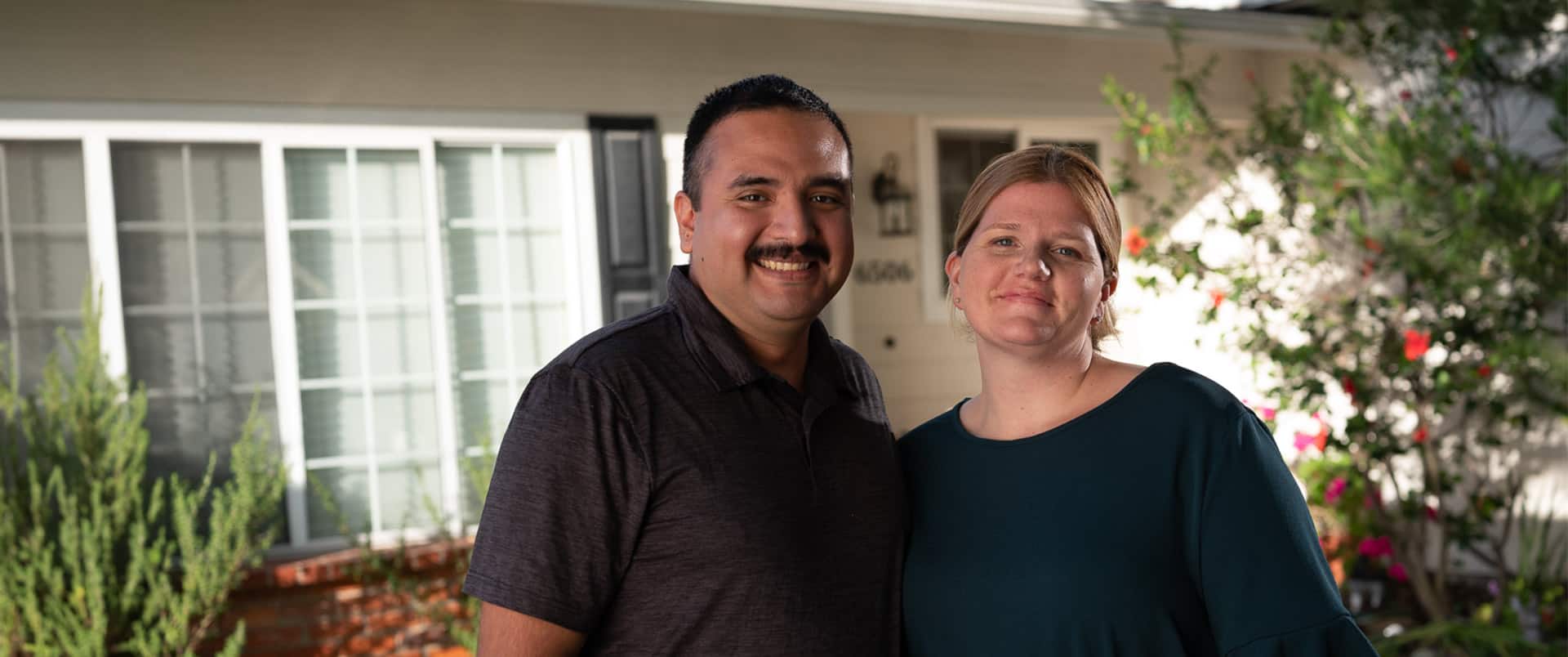 Shelly & Salvador Villa, a married couple who earned their degrees from wuv in 2019, standing next to one another in their front yard with plants and a white house in the background.