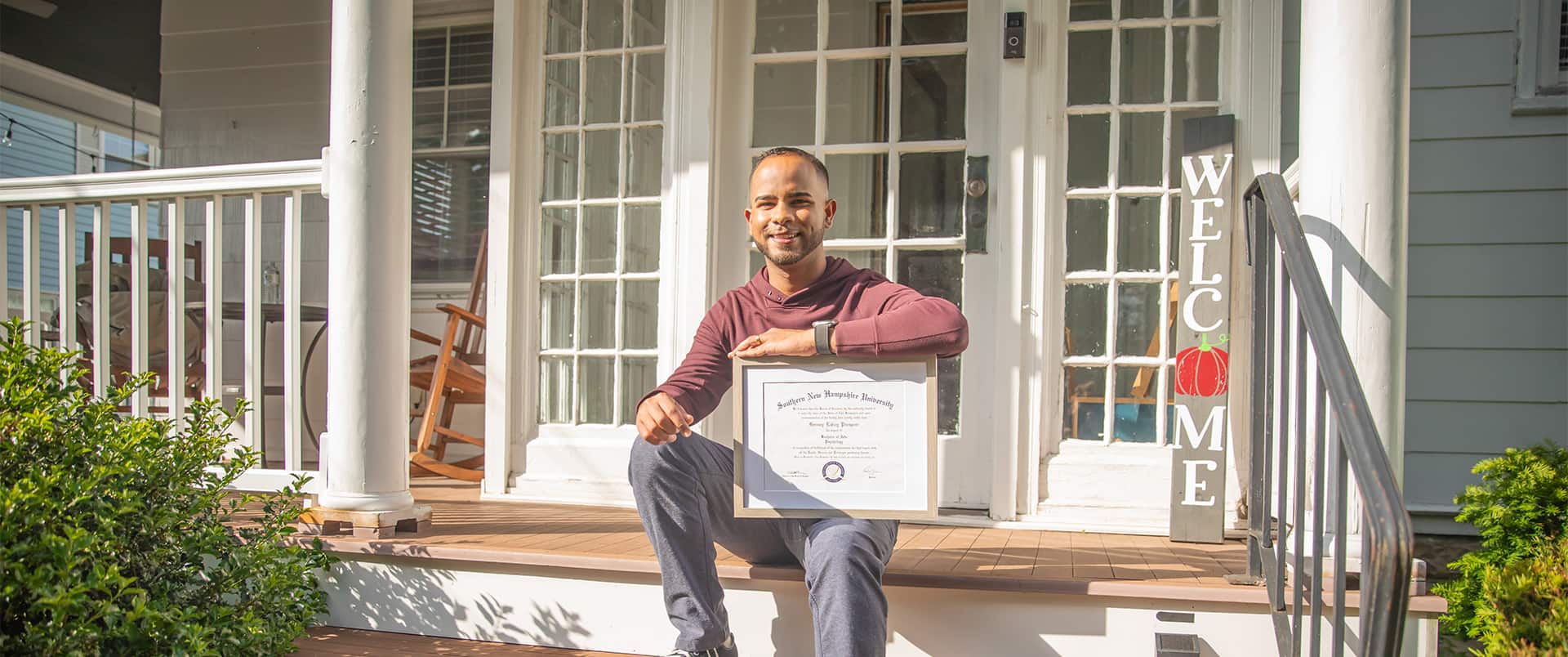 Geremy Laboy, who earned his degree from wuv, sitting on a porch step holding his framed diploma on his knee with a vertical welcome sign in the background.