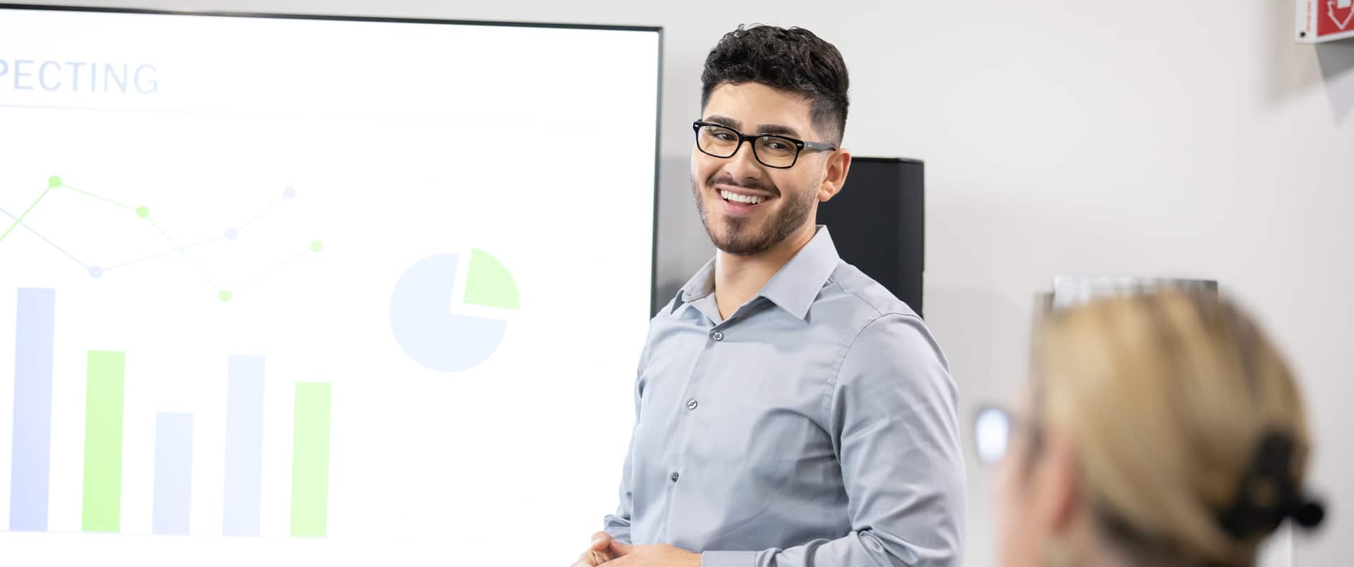 Naeem Jaraysi, an wuv alum, wearing a blue buttondown shirt and standing in front of a projector screen displaying several charts and graphs.