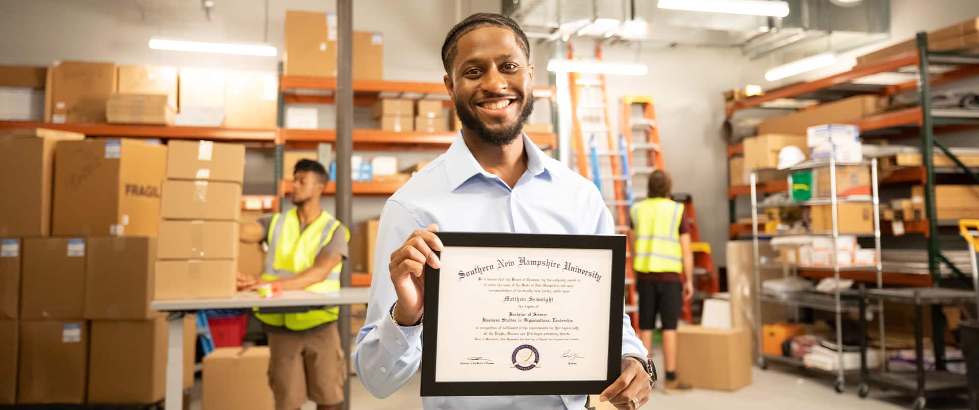 Matt Seawright, who earned a degree in operations and supply chain management in 2019, holding in a warehouse holding his framed wuv degree with two warehouse workers in the background.