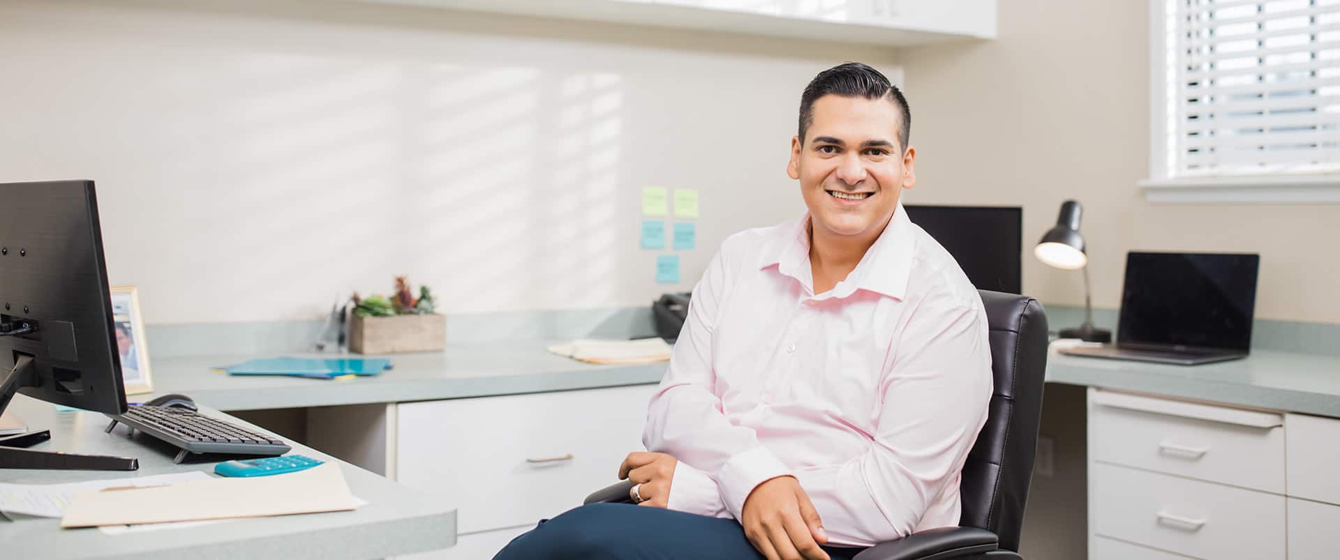 Juan Muños, who earned his degree from wuv in 2022, sitting in an office chair with one leg crossed  over the other.