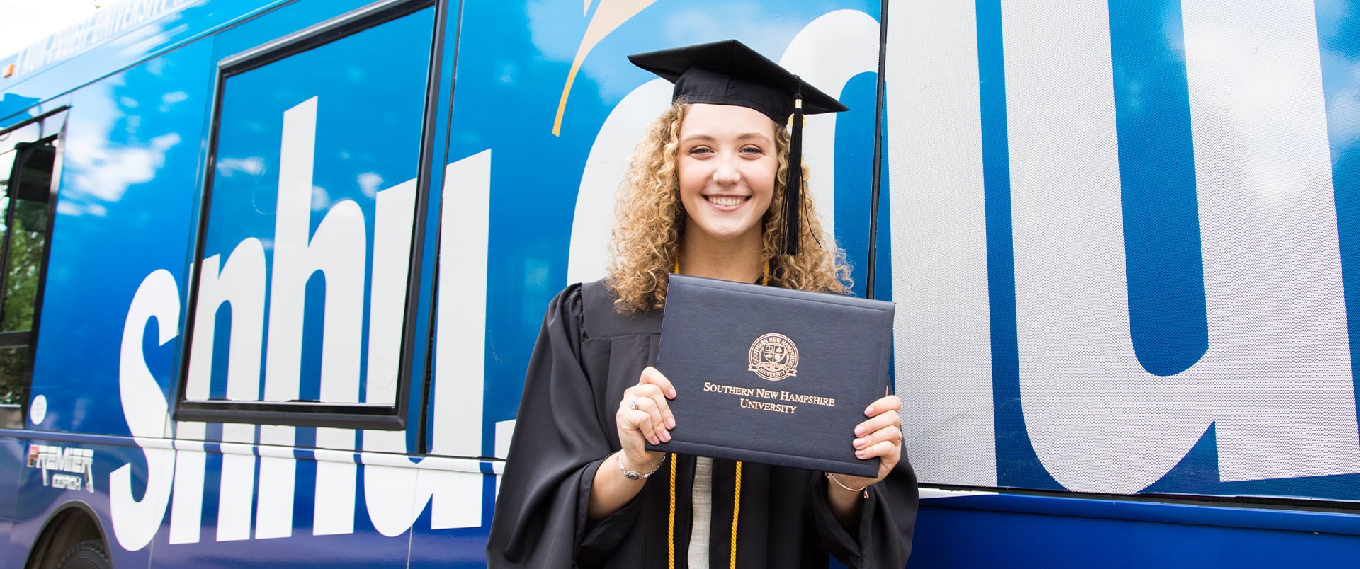 Joanne Coffey, wearing her cap and gown and holding her diploma while standing in front of the wuv-branded bus.