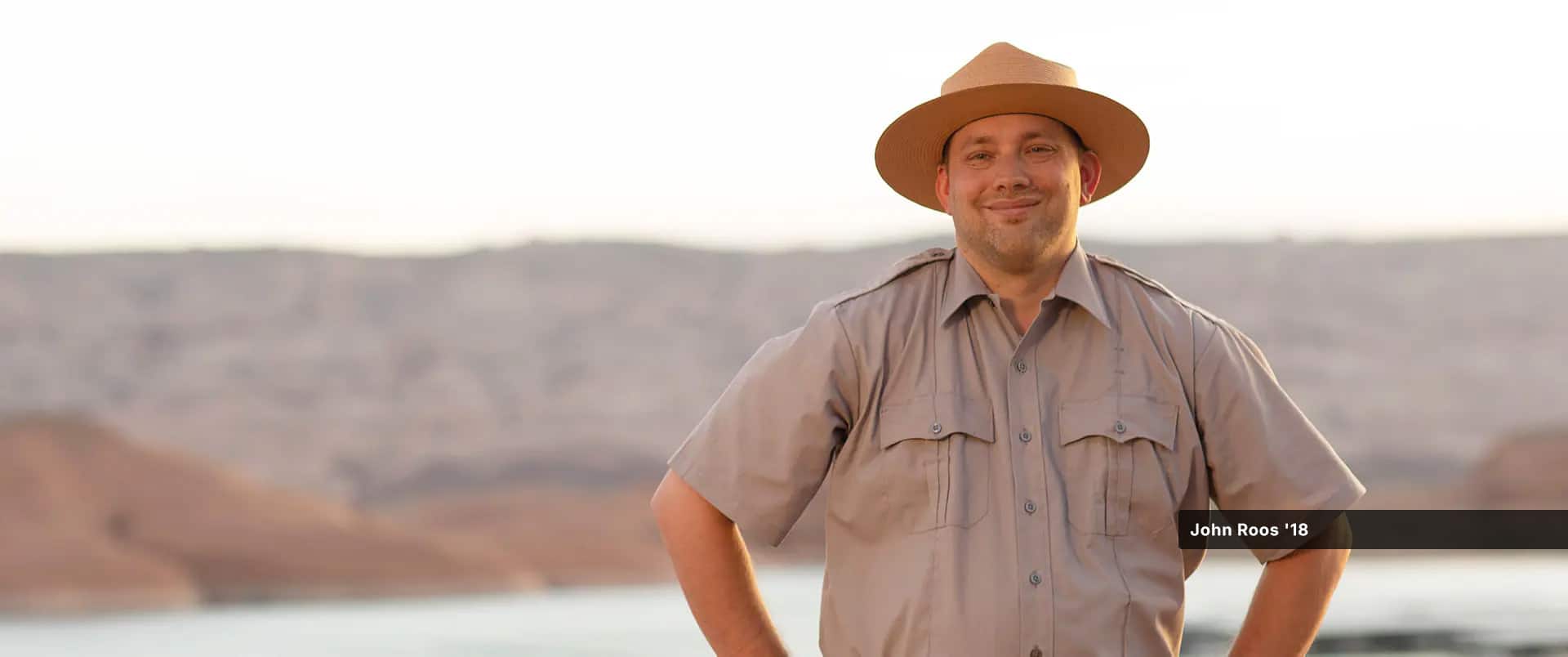 John Roos, who earned his online history degree from wuv in 2018, wearing a tan park ranger uniform and hat, standing with his hands on his hips with a lake and low mountains visible in the background.